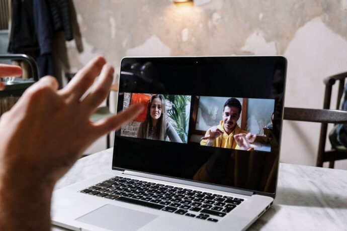 Laptop med videomøde, hvor to deltagere ses, og en hånd vinker foran skærmen